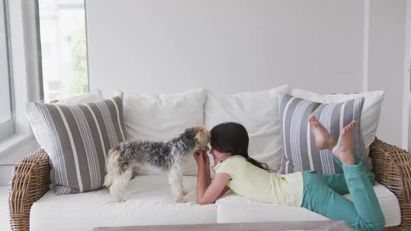 Caucasian girl spending time at home, lying on sofa with face mask, playing with pet dog, in slow mo