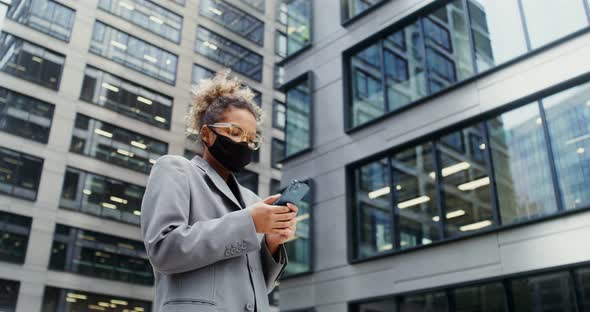 A Business Woman in a Medical Mask is Typing on a Mobile Phone Outdoors