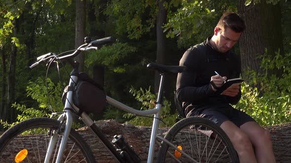 A Young Handsome Cyclist Sits on a Log Next To His Bike in a Forest and Writes Notes