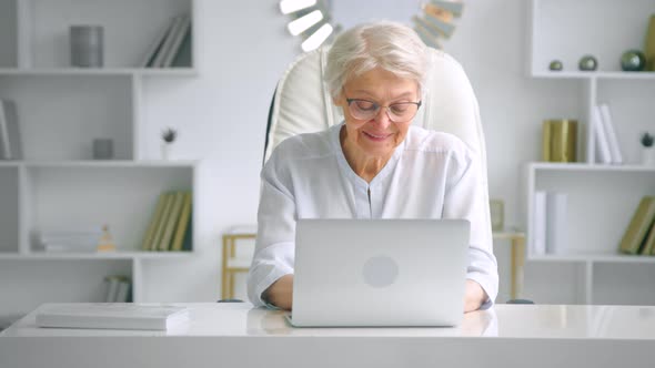 Smiling aged woman in glasses looks at laptop keyboard