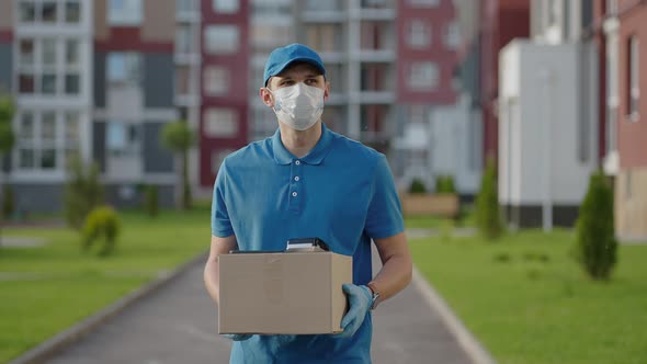 The Male Deliveryman in a Cap and a Protective Mask and Gloves Goes with a Box in His Hands and