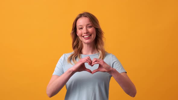 Smiling blonde woman wearing blue t-shirt showing heart shape gesture at the camera