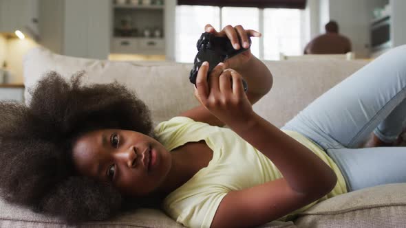 African american daughter lying on couch playing video game with father in background