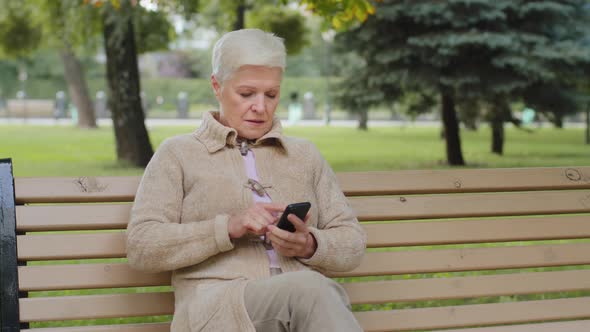 Attractive Grayhaired Senior Woman of Retirement Age Sitting on Park Bench Making Video Call Looking