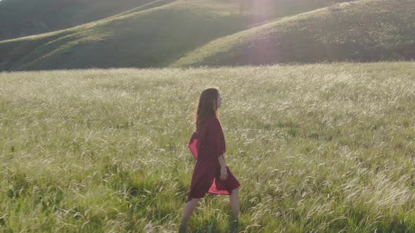 Aerial View of a Young Woman in a Red Dress Walking Barefoot Along the Hillside in High Flowering