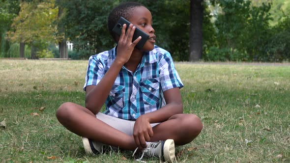 A Young  Black Boy Sits on Grass in a Park and Talks on a Smartphone
