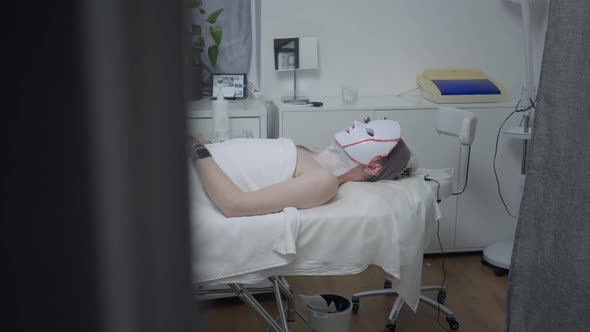 Side View Wide Shot of Relaxed Tattooed Male Client Lying on Beauty Bed in Spa with Innovative Red