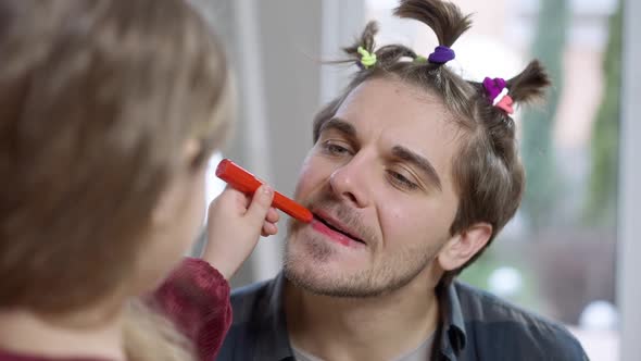 Headshot of Joyful Caucasian Young Man Laughing As Daughter Painting Parent Lips with Red Marker