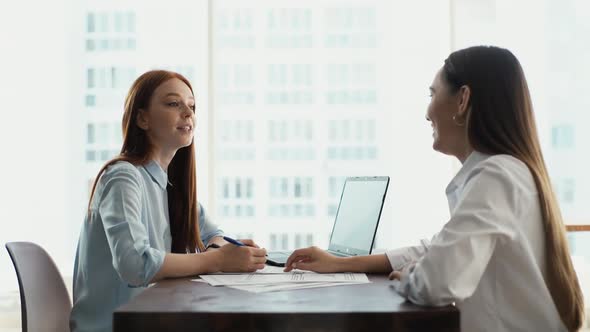 Side View of Two Females Colleagues Discuss Financial Data in Report Using Laptop and Paper