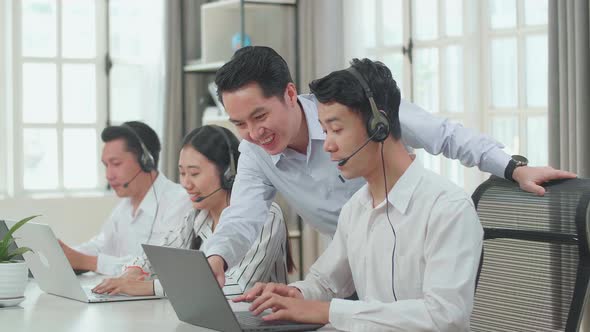 A Man Coming To Help One Man Of Three Asian Call Centre Agents In Headsets To Work With Computer