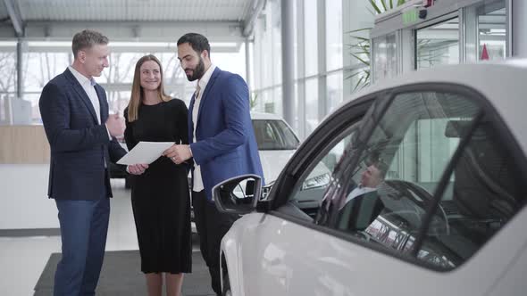 Young Rich Multiracial Family Buying Car in Dealership. Smiling Middle Eastern Client and Caucasian