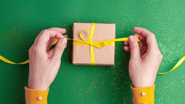Human hands tied and untied brown gift box with a yellow satin ribbon bow on jade green background