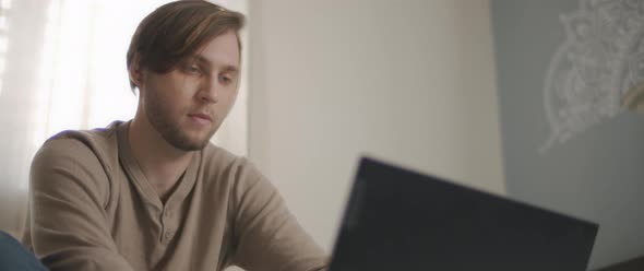 A close up of a young man sitting in bed and working on a computer. 