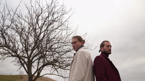 Two Men in Coats Stand with Their Backs to Each Other Near a Dried Tree