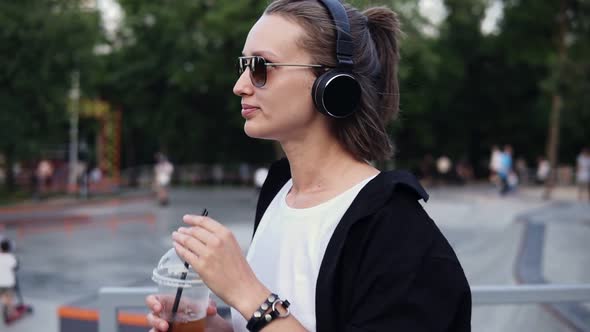 Side View of Aucasian Girl Walking Through Park Side and Drinking a Soft Drink with a Straw