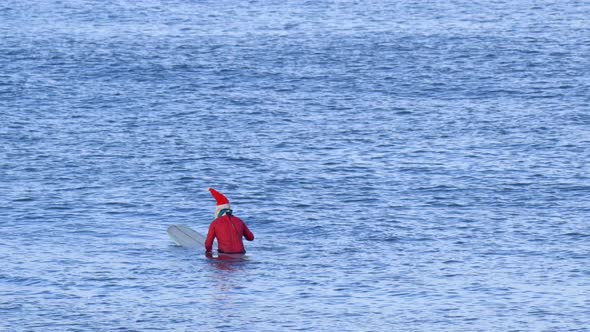 Santa Claus sits on his surfboarding waiting for waves while surfing.