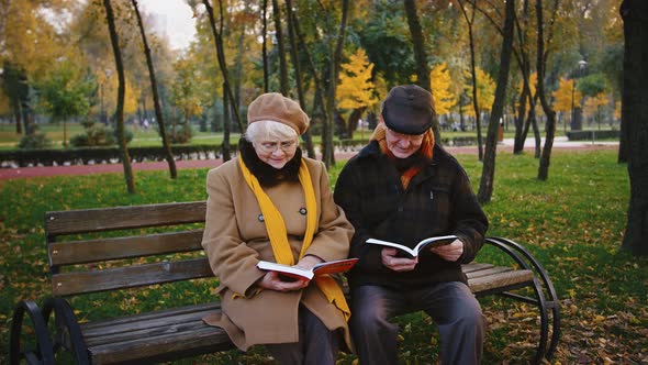 Retired Couple in Glasses and Warm Outerwear are Reading Books While Sitting on Wooden Bench in