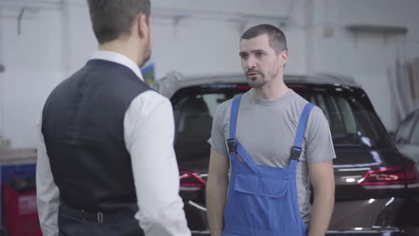 Young Caucasian Man in Workrobe Taking Customer's Car Keys in Repair Shop and Shaking His Hand. Auto