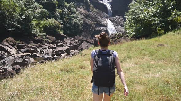 Woman Tourist Backpack Jogs Path To Rushing Waterfall Raises Hands Up