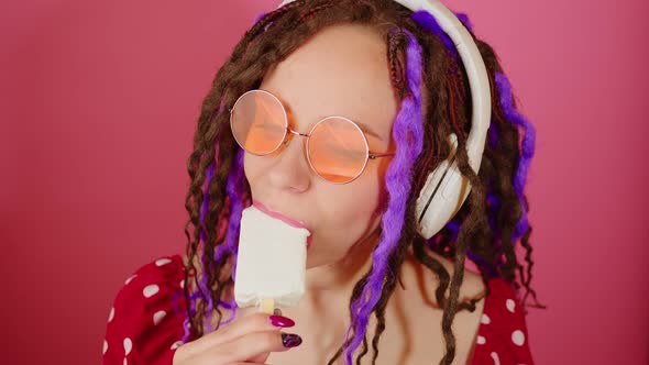 Young beautiful woman in bright sunglasses, wireless white headphones licking ice cream