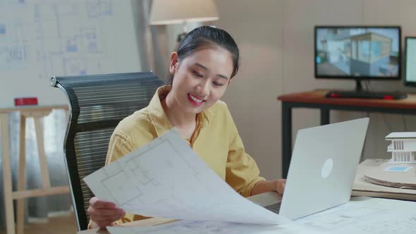 Asian Woman Engineer Looking At Blueprint And Typing On A Laptop At The Office