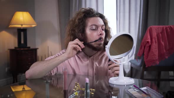 Front View of Young Caucasian Man Looking at Mirror and Applying Mascara on Mustaches. Intersex