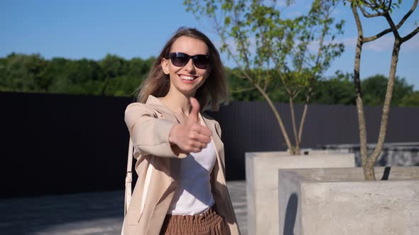 Positive Ambitious Young Woman in Sunglasses is Showing Thumbs Up Outdoor