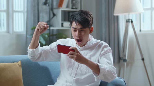 Asian Man Celebrating Winning Game On Smartphone While Lying On Sofa In The Living Room