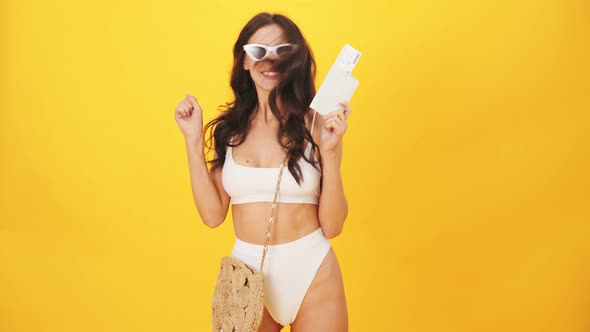 A cheerful brunette woman wearing a swimsuit jumping while holding boarding pass tickets and passpor