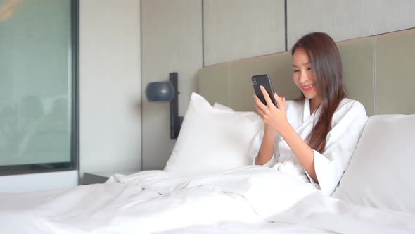 Smiling Asian Woman in Bedroom Checking News, Messages on Smartphone on a Bright Morning With Yes Ha