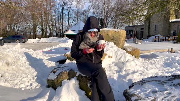 Woman Sits On The Snow Holding A Cellphone And A Cup On A Sunny Day. wide shot