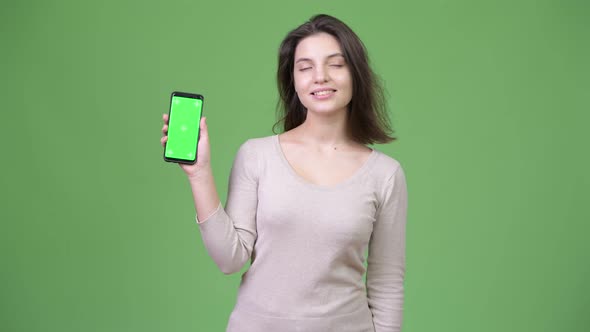 Young Happy Beautiful Woman Thinking While Showing Phone Against Green Background