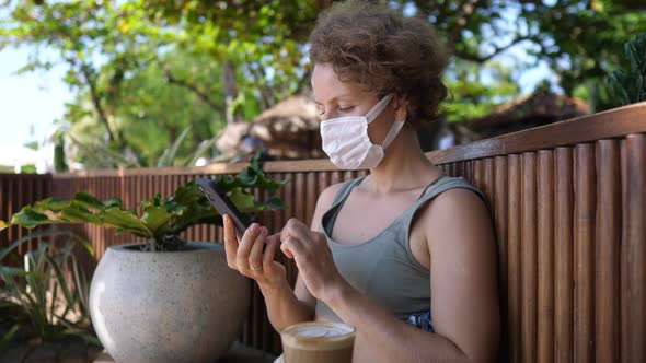 Young Woman in a Face Mask Relaxing in an Tropical Garden Drinking Latte Coffee and Browsing on Her