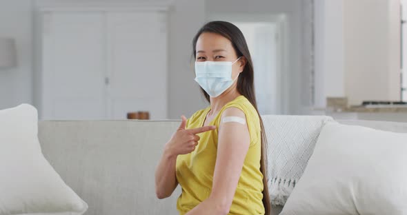 Happy asian woman in face mask sitting on sofa showing arm with plaster after vaccination