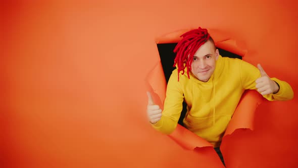 Young Handsome Man Showing Thumbs Up and Sticking Out of Hole of Orange Background