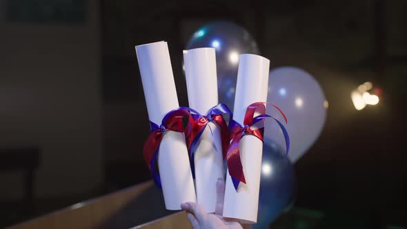 Certificate of Graduation Tied with Red and Blue Ribbon in Hand Background of Balloons and Light