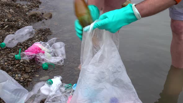 A man in rubber gloves collects plastic waste in a river in a plastic bag