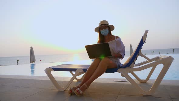 Young Woman in Protective Mask, Sun Hat and Summer Clothes, Working on Laptop, Sitting on Sun