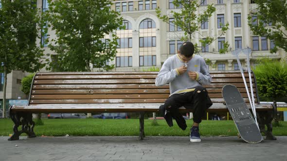 Attractive Young Man Sitting on the Bench in the Park Listening To Music on His Cellphone Then