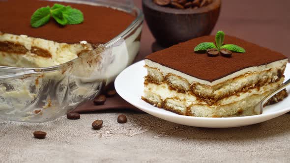 Portion of Traditional Italian Tiramisu dessert and baking dish on grey concrete background