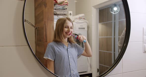 A Teenage Girl Brushes Her Teeth with an Electric Toothbrush in the Bathroom