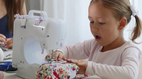 A Little Girl Is Sewing Fabric and Talking. Mom Is at the Background. Close-up.