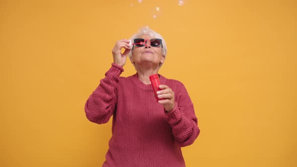 Trendy Senior Woman with Sunglasses Blowing Soap Bubbles. Isolated on Orange Background