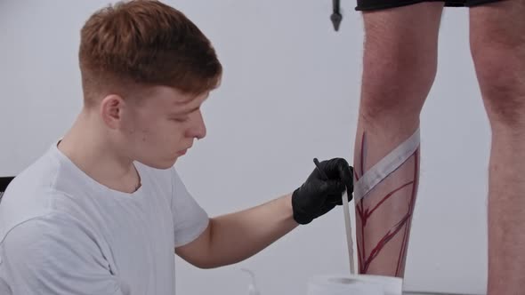 Young Man Tattoo Artist Taping a Duct Tape on the Leg on His Client in the Salon