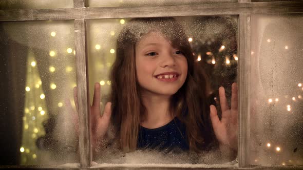 Cute girl looking through winter window at Christmas time