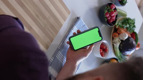 Man using a Green Screen Display Smartphone in the Kitchen, Vegetables in Background