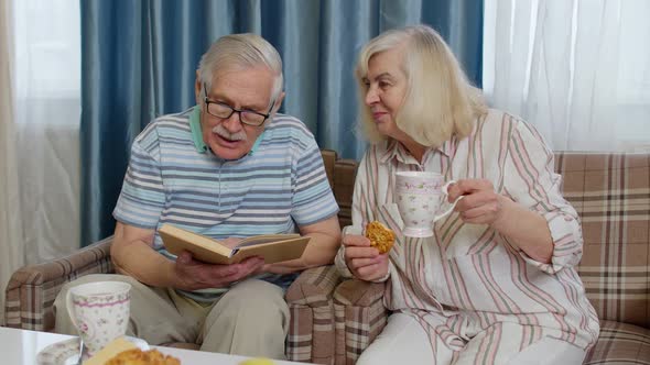 Smiling Senior Couple Grandfather Grandmother Resting on Sofa Drinking Coffee Reading Book at Home