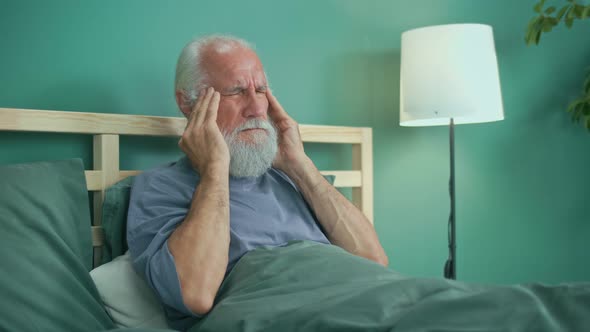 Tired Elderly Man Has Acute Headache While Sitting on Ahe Bed At Home