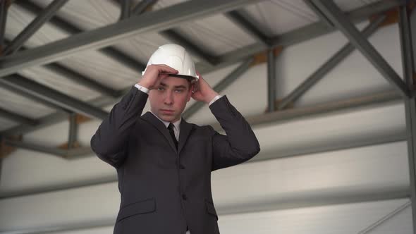 A Young Man Puts on a Protective Helmet and Shows a Thumb at a Construction Site. The Boss in a Suit