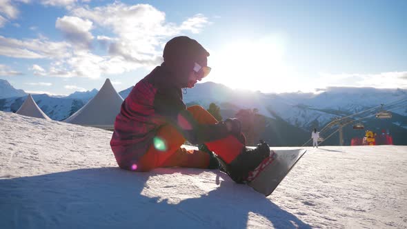 Snowboarder Girl Sitting on Relax Moment at Sunset in Ski Resort. Girl Snowboarder Enjoys the Ski